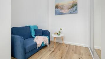 Quarto com um sofá azul, uma mesinha branca com uma planta e um quadro de uma praia na parede.