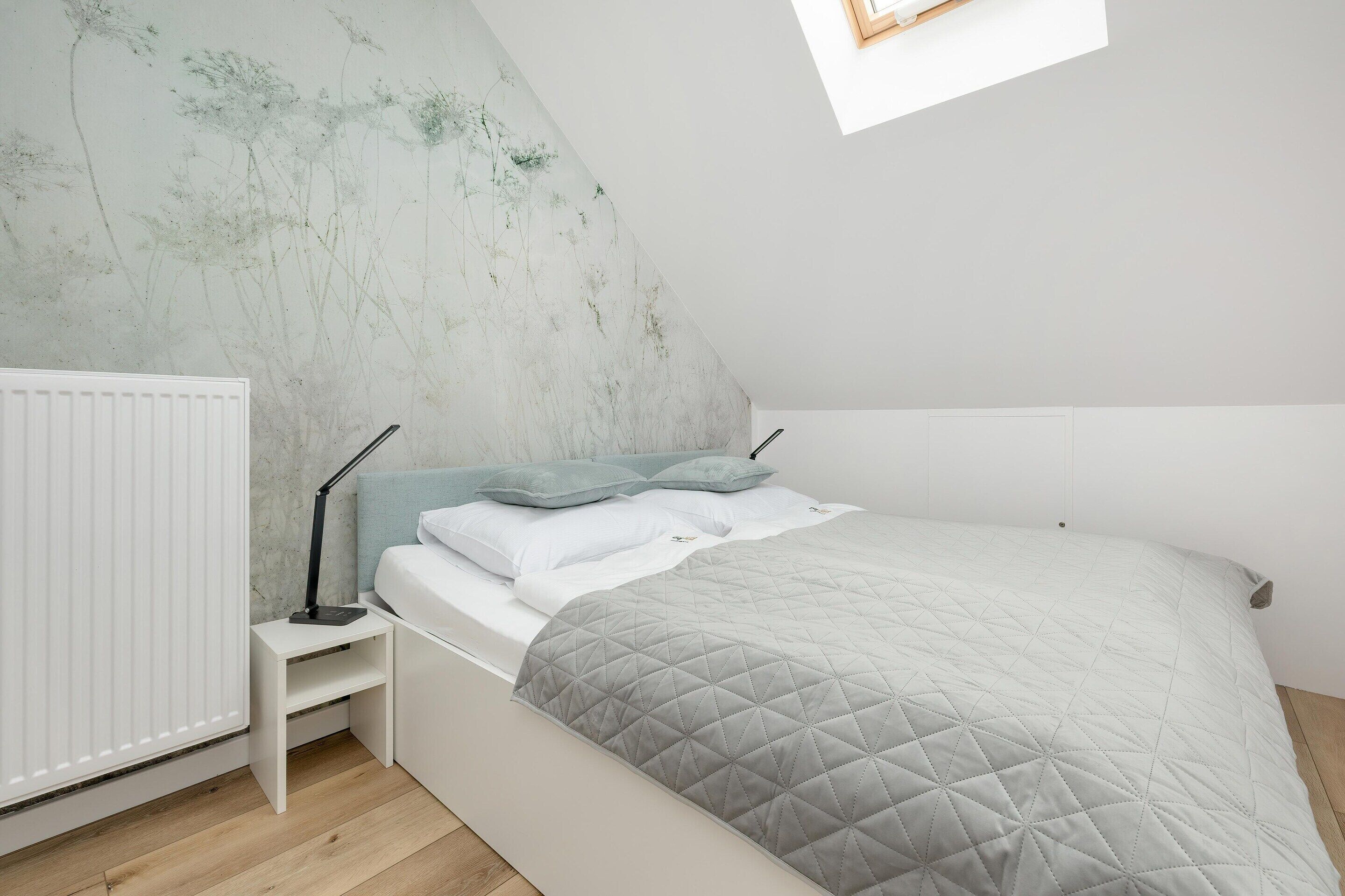 Une chambre avec un plafond en pente, un lit recouvert d’une couette grise matelassée, une table de chevet avec une lampe et un radiateur.