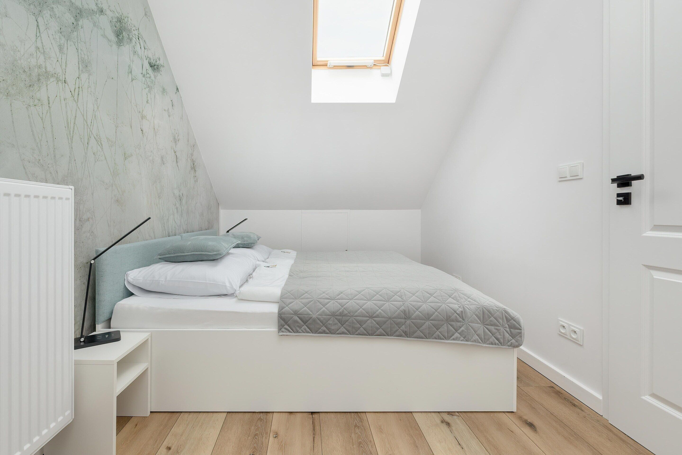 A compact, modern bedroom with a skylight, a built-in bed, a bedside table, and a radiator.
