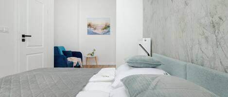 A bedroom with a bed, a chair, a small table with a plant, and a painting on the wall.