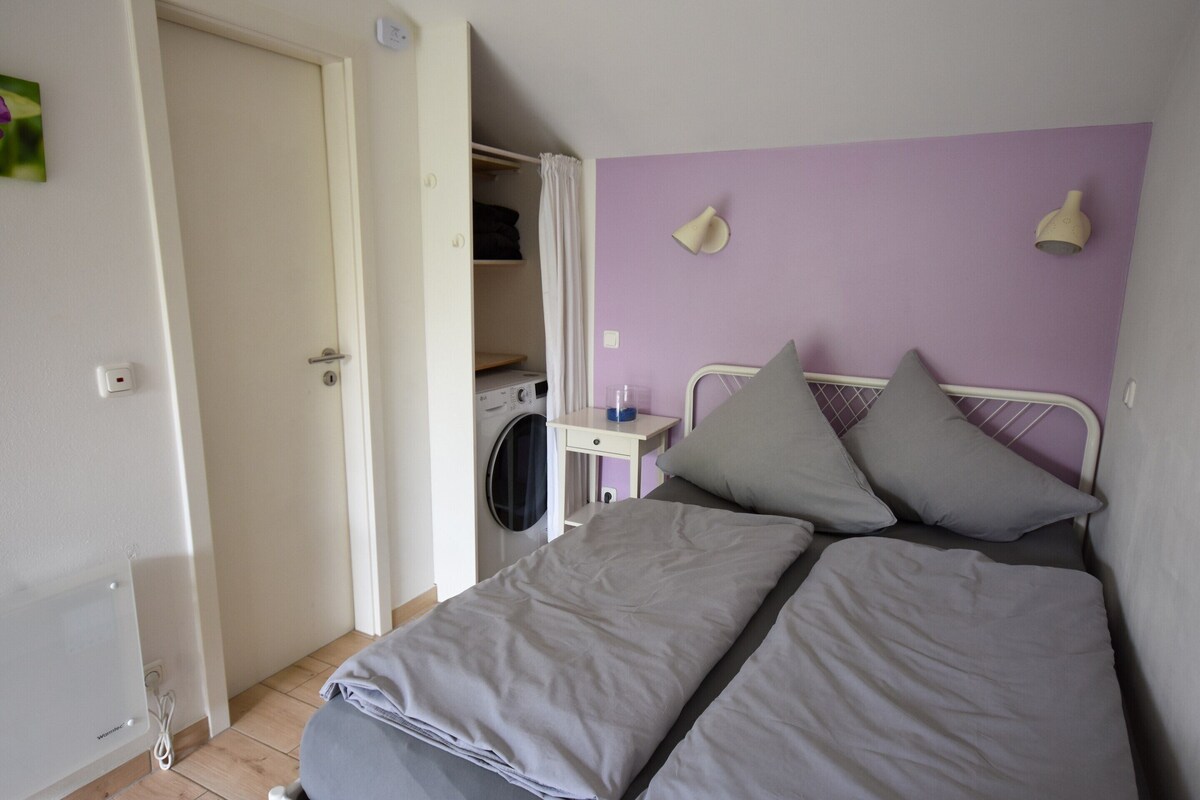 A bedroom with a bed, a washing machine, a small table, and a closet.