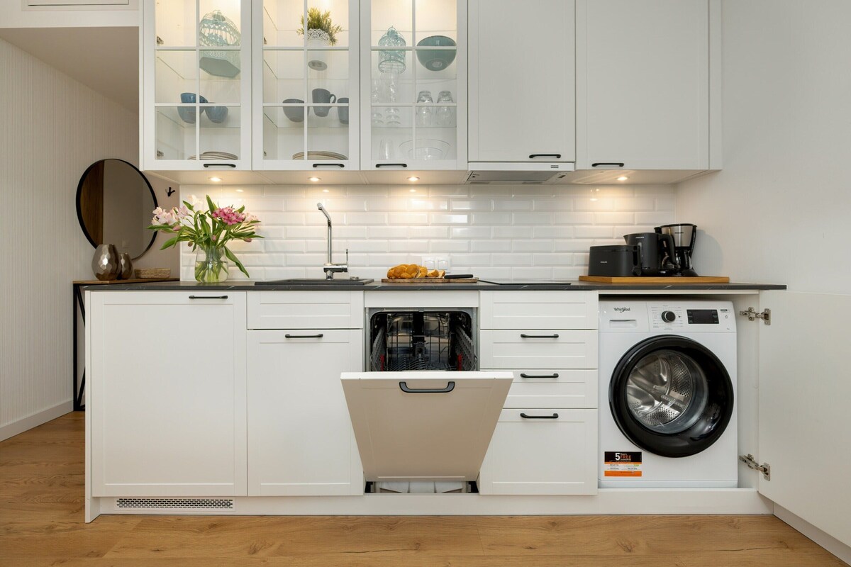 A compact kitchen with white cabinetry, a built-in dishwasher, a washing machine, and a small countertop with a coffee maker.