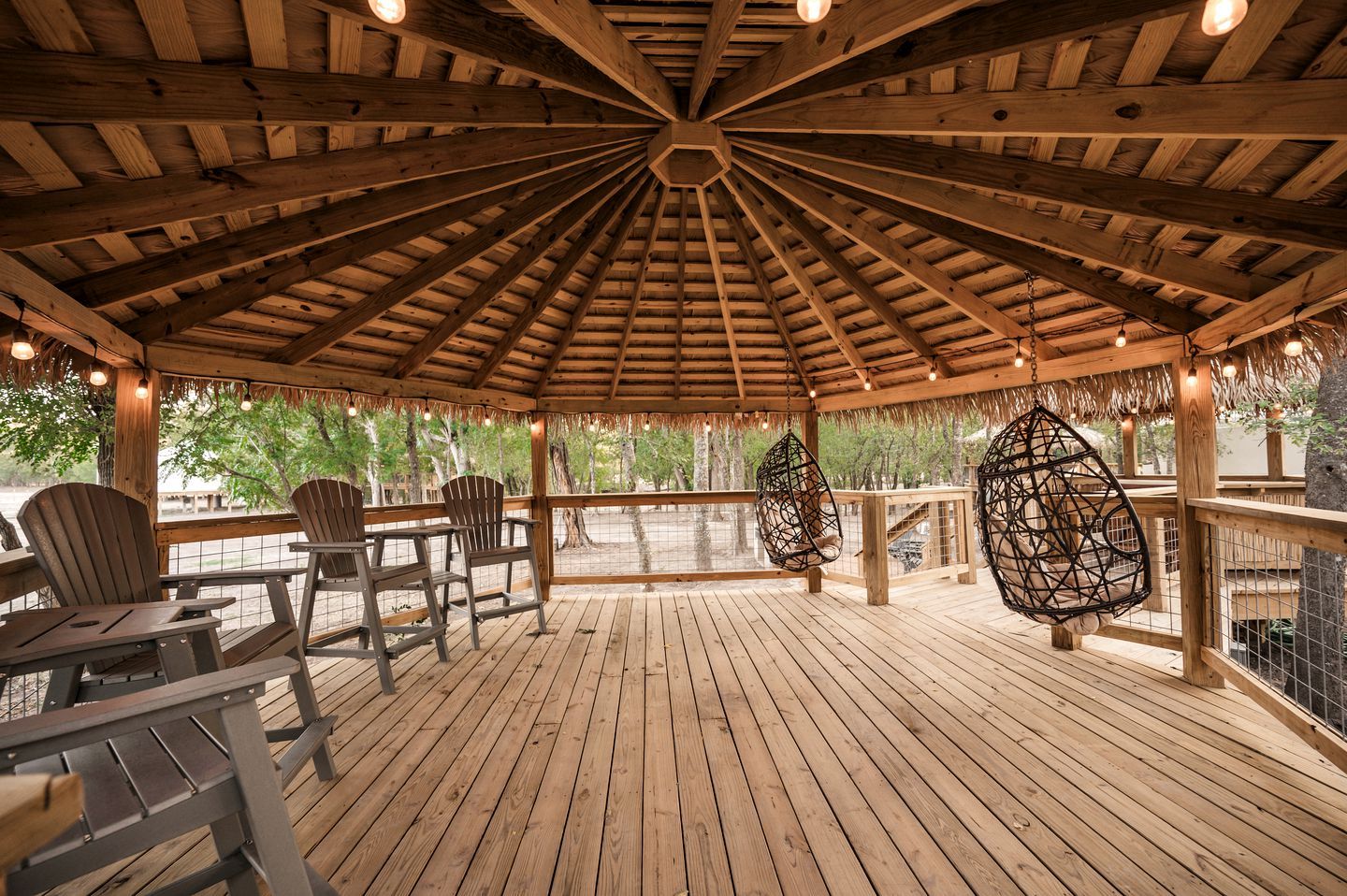 A wooden gazebo with a thatched roof, seating area, and hanging chairs.