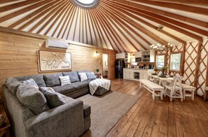 A spacious living area with a large sofa, a dining table with chairs, a kitchen area, and a wooden ceiling structure. - A Yurt for Everyone: Family and Friends Getaway in Bandera, TX (Bandera)