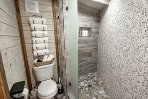 A bathroom with a wooden floor, a white toilet, a towel rack, and a shower area with a textured wall. - A Yurt for Everyone: Family and Friends Getaway in Bandera, TX (Bandera)