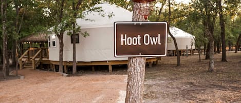 Un letrero con la leyenda 'Hoot Owl' sujeto a un árbol, con una gran carpa blanca y estructuras de madera al fondo.