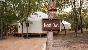 A sign reading 'Hoot Owl' attached to a tree, with a large white tent and wooden structures in the background. - A Yurt for Everyone: Family and Friends Getaway in Bandera, TX (Bandera)