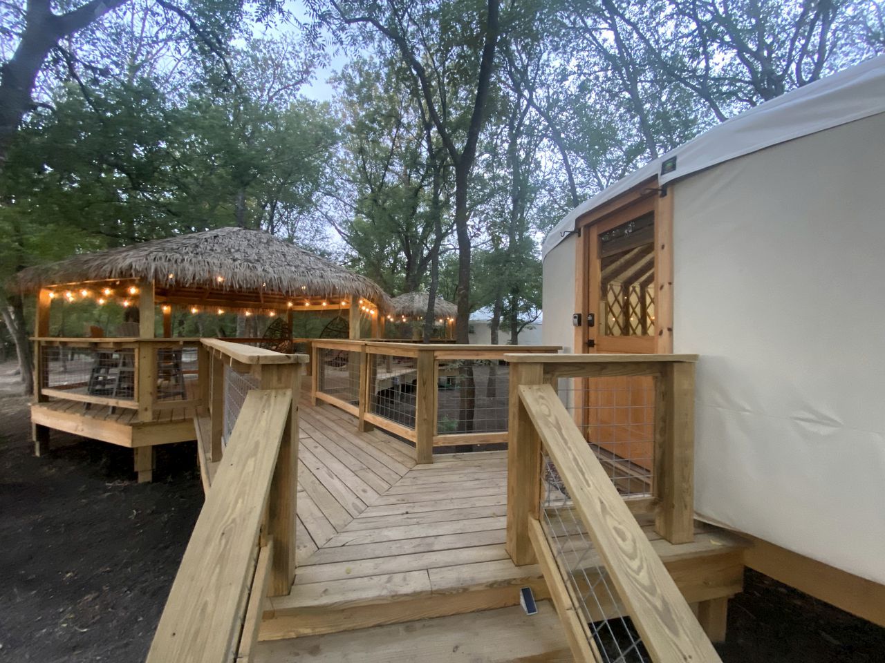 Eine Holzterrasse führt zu einem Gebäude mit einem Strohdach, Lichterketten, einer weißen, zeltartigen Struktur und einer Holztür.