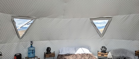 Una habitación con carpa que incluye una cama, dos ventanas, un ventilador y una botella de agua.