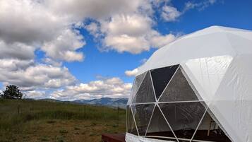 A geodesic dome structure with a clear view of the surrounding landscape, including grass, a fence, and distant mountains.