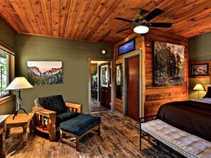 A room with wooden flooring, a ceiling fan, a bed, a chair, a table, and a painting on the wall.