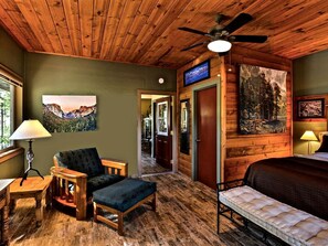 A room with wooden flooring, a ceiling fan, a bed, a chair, a table, and a painting on the wall.