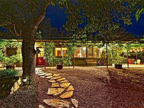 A well-lit outdoor area with a stone pathway leading to a covered seating area, surrounded by trees and potted plants.