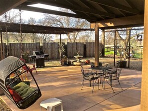 A covered patio with a swing chair, a grill, a dining table with chairs, and a fountain.