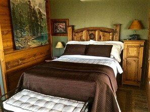 Een houten bed met hoofdeinde, een nachtkastje met lamp, een schilderij aan de muur, en een houten kast.