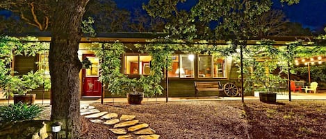 A well-lit outdoor area with a stone pathway leading to a covered seating area, surrounded by trees and potted plants.