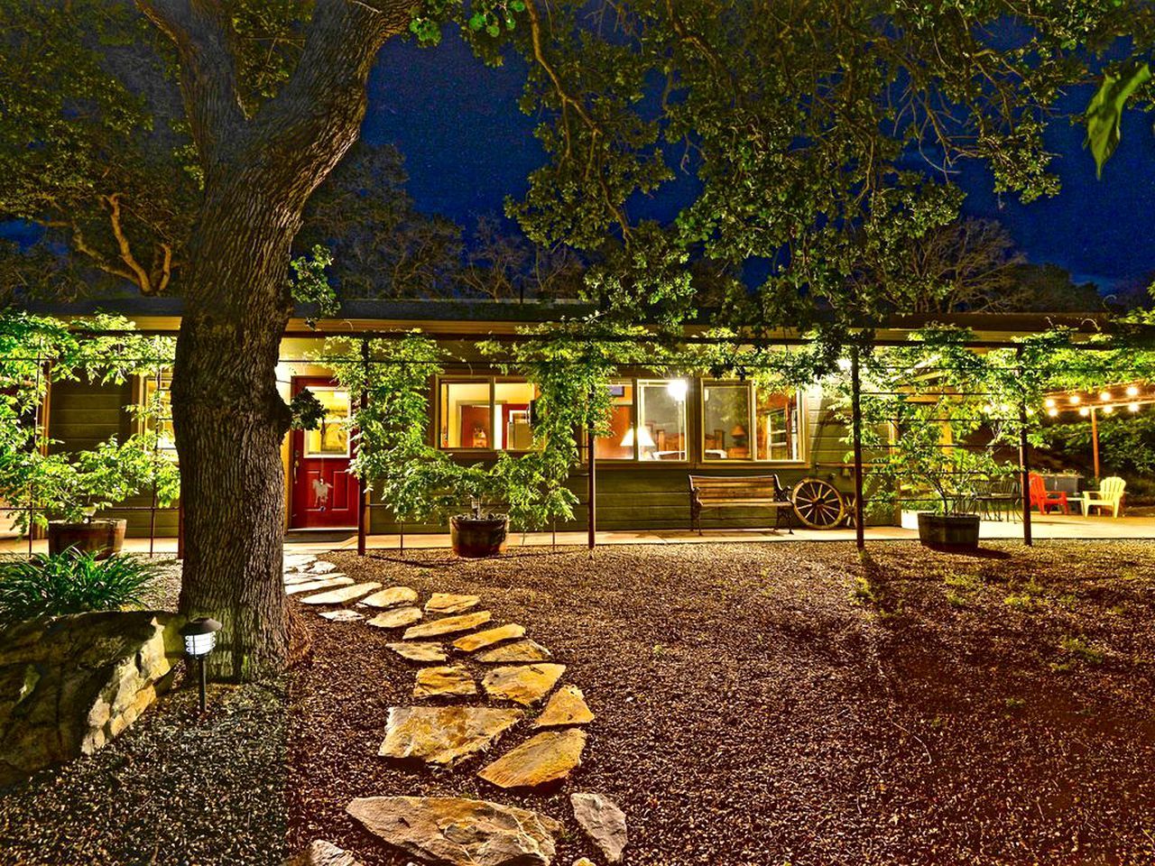 A well-lit outdoor area with a stone pathway leading to a covered seating area, surrounded by trees and potted plants.