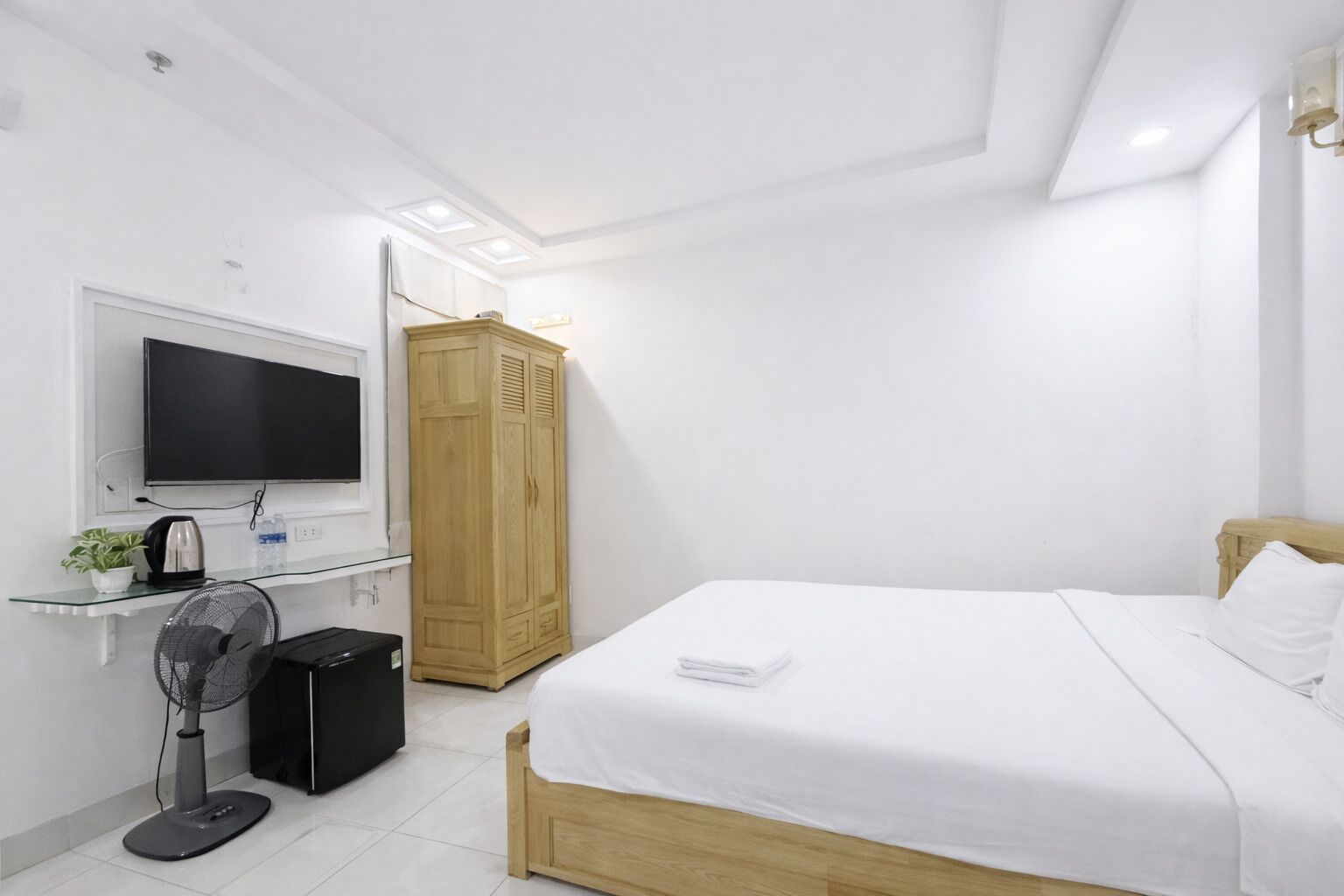A bedroom with a bed, a TV, a wardrobe, a desk with a fan, and a small plant.