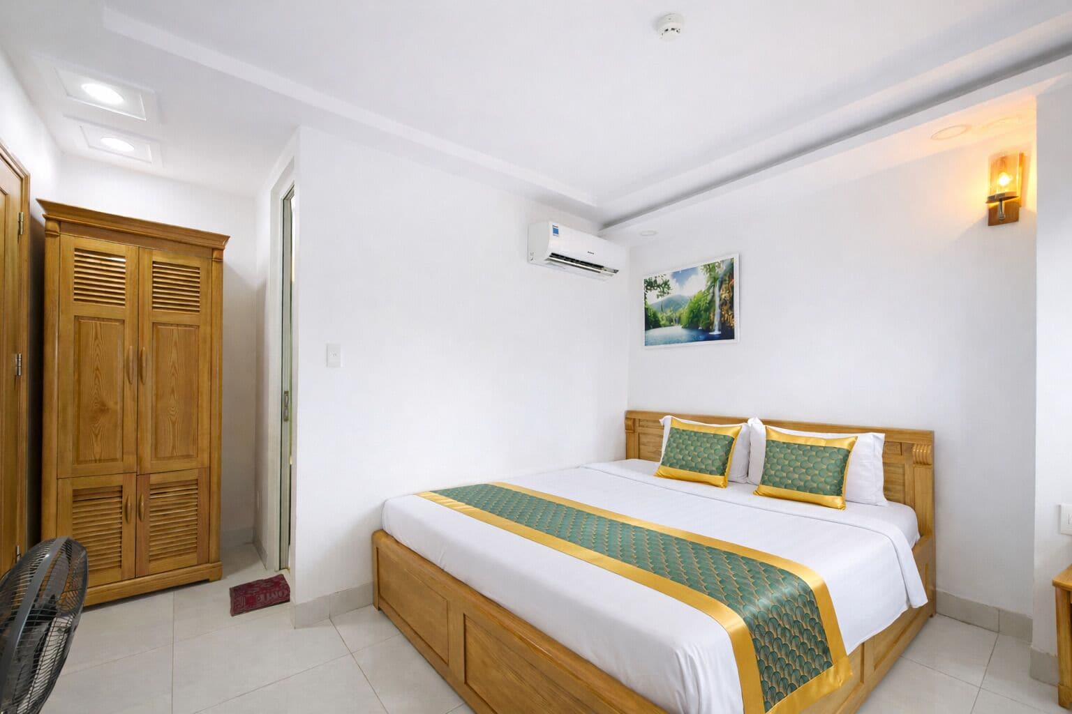 A hotel room with a bed, wooden wardrobe, air conditioning unit, and a painting on the wall.