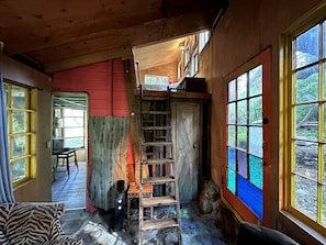 L’intérieur d’un chalet en bois, avec une échelle en bois, un escalier et une vue sur l’extérieur grâce à de grandes fenêtres.