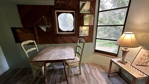 Et spiseområde med et træbord og stole, et vindue med udsigt til grønne omgivelser, og en lampe på et sengebord.
