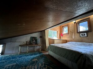 Un dormitorio con techo inclinado, una cama, una mesita con objetos y un ventanal con vistas.