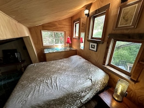 A bedroom with a bed, a desk, and a window with a view of greenery.