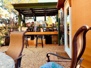 Un patio exterior con mesa y sillas de madera, una parrilla y una zona cubierta con una pérgola.