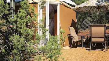 A small, single-story structure with a door open, surrounded by sparse vegetation and a table with chairs.