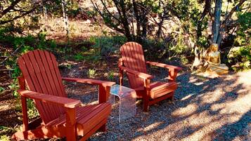 Two wooden Adirondack chairs and a small table in a shaded outdoor area with gravel ground.