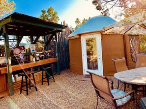 Un área de cocina rústica al aire libre con barra de madera, taburetes y un espacio de estar techado.