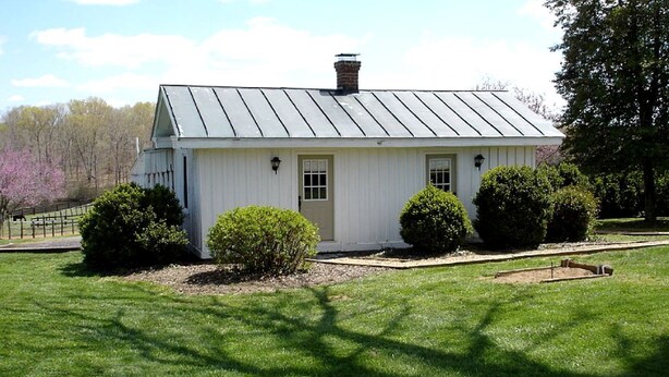 A white single-story building with a metal roof, a front door, and two small bushes. - Pre-Civil War Servants' Cottage on Huge Horse and Cattle Farm near Charlottesville, Virginia (Gordonsville)