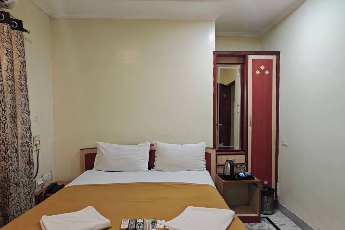 A hotel room with a bed, pillows, a bedside table with a kettle, a mirror, and a trash bin.