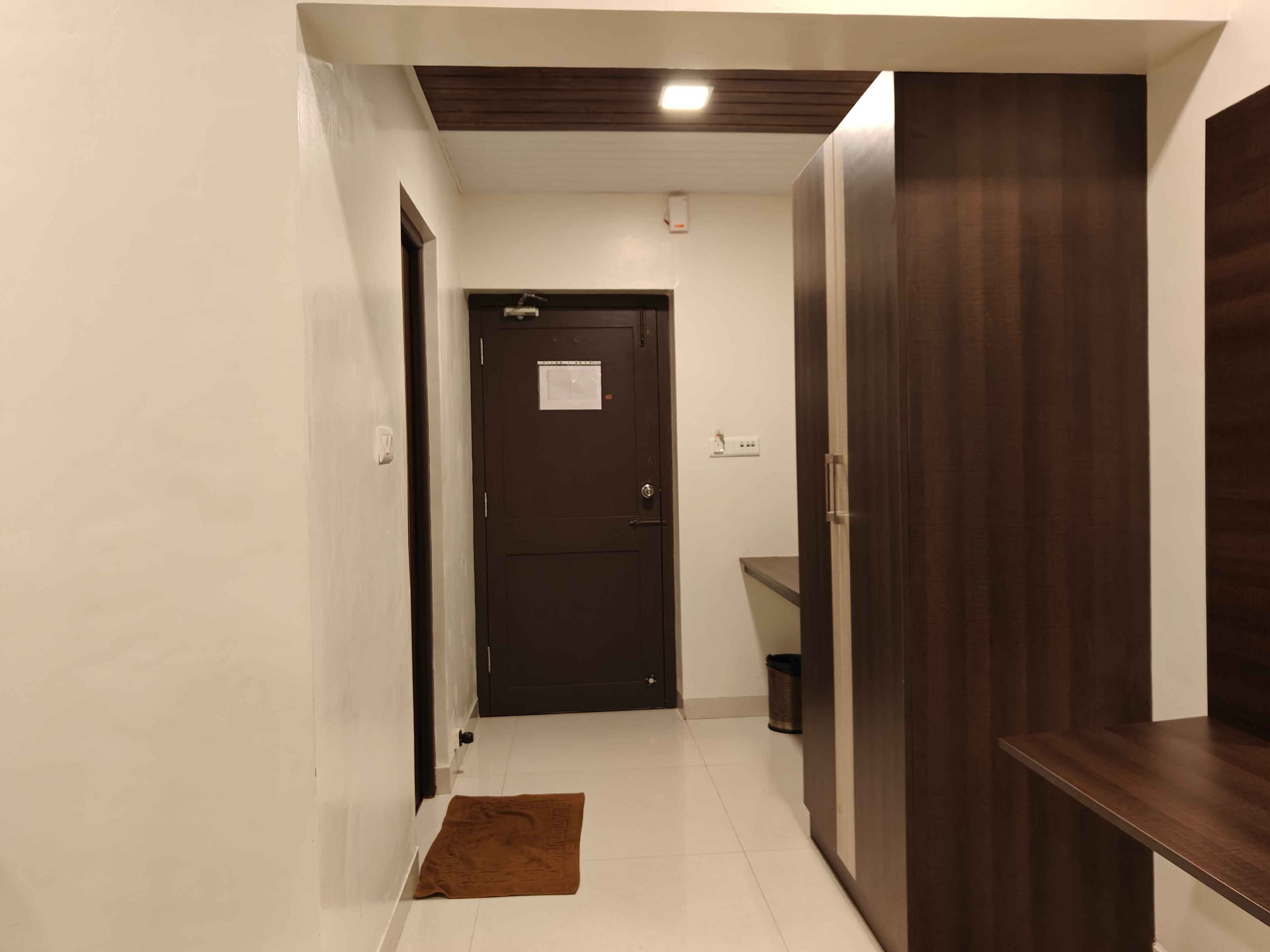 A hallway with a dark wooden door, a white wall, a tiled floor, and a brown mat.