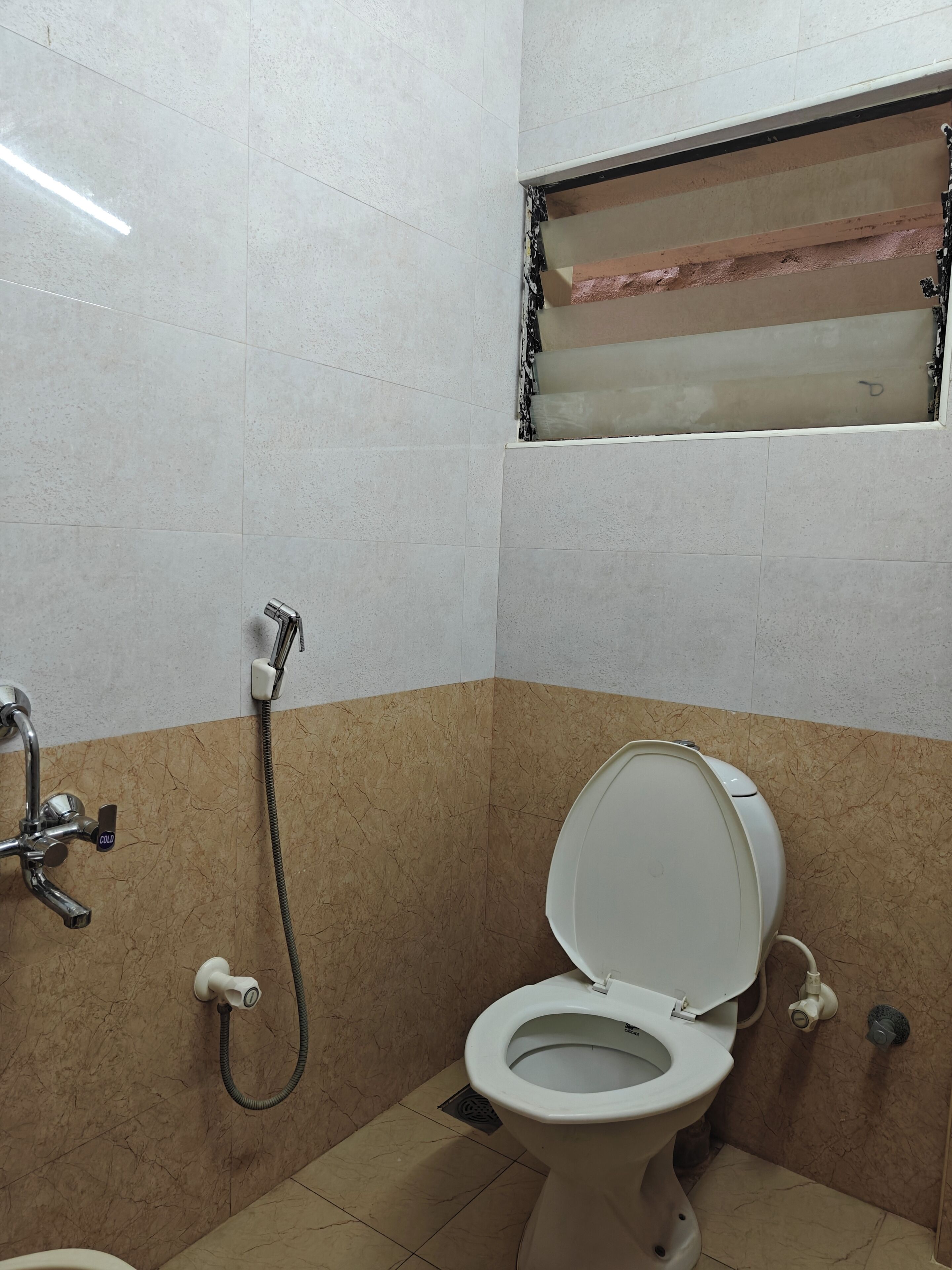 A bathroom with a toilet, shower, and a visible window with a damaged frame.