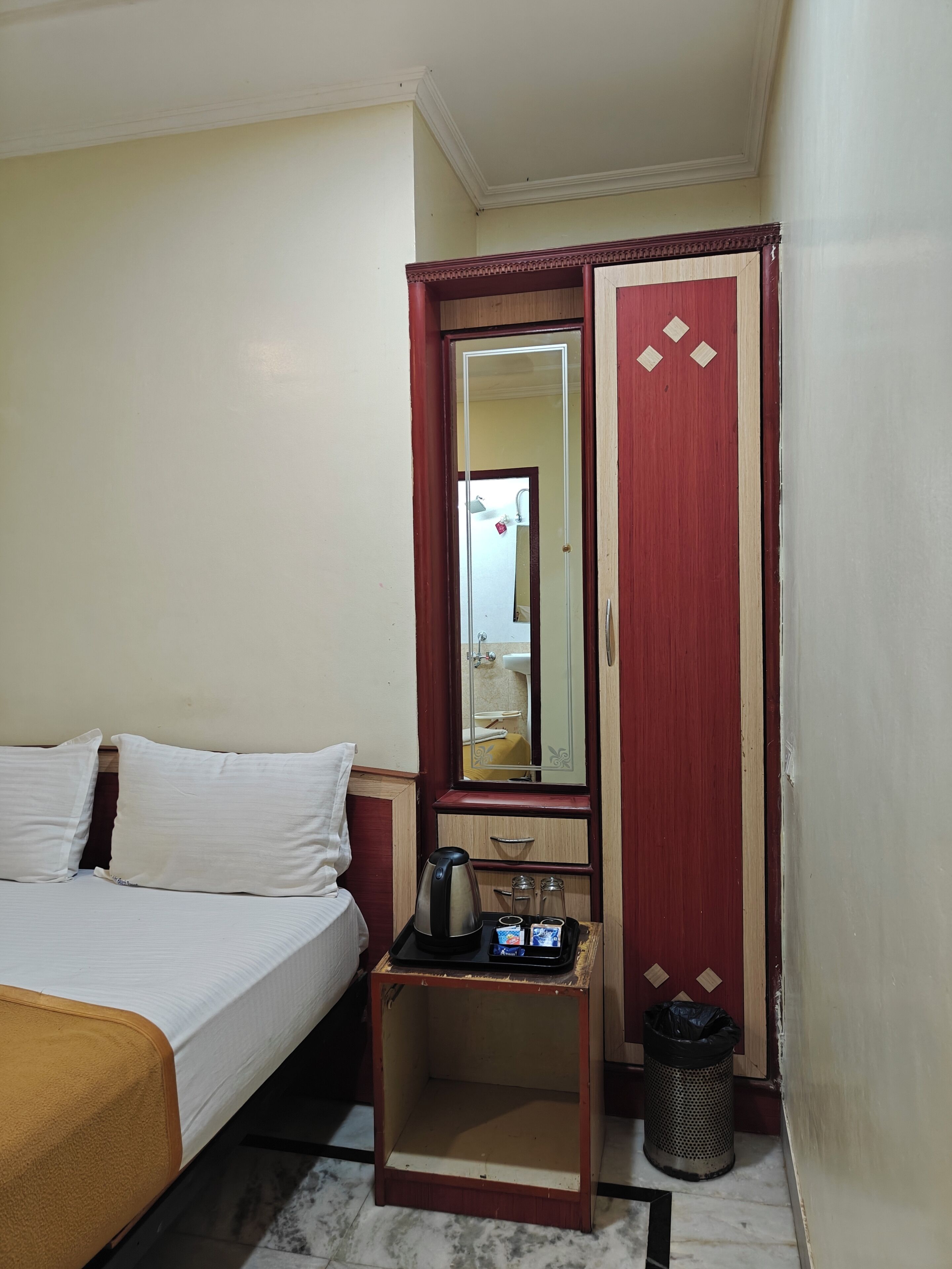 A hotel room with a bed, a small table with a kettle, a mirror, and a wardrobe.