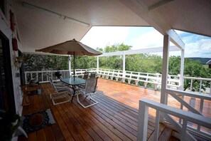 A wooden deck with a table and chairs, a white railing, and a view of greenery. - Riverfront Cabin with Spacious Interior, Large Deck and Views in Texas (Wimberley)