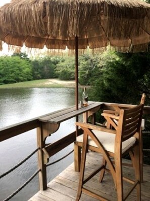 A wooden deck with a thatched umbrella, a wooden chair, and a view of a body of water. - Texas Safari Camp Hummingbird Tree House (Kaufman)