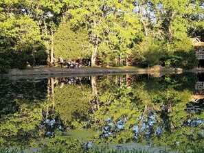 A serene lake surrounded by dense trees, a wooden dock, and a small structure on stilts. - Texas Safari Camp Hummingbird Tree House (Kaufman)
