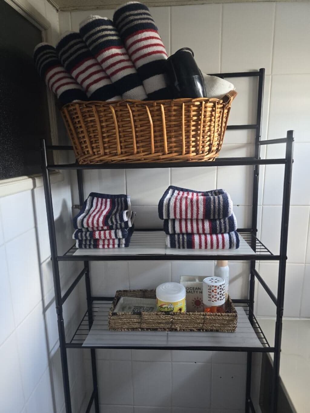 A metal shelf unit with a wicker basket on the top shelf, folded blankets on the middle shelf, and a tray with items on the bottom shelf.