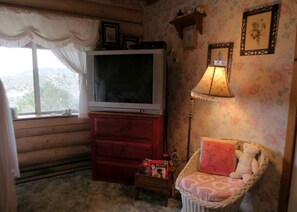 Ein gemütliches Zimmer mit Fernseher auf einer Holzkassette, ein Korbstuhl mit Kissen, ein kleiner Tisch mit einer Schachtel und einer Lampe.