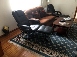 Una habitación con un sofá marrón, dos sillas de oficina negras, una mesita de madera y una alfombra estampada.