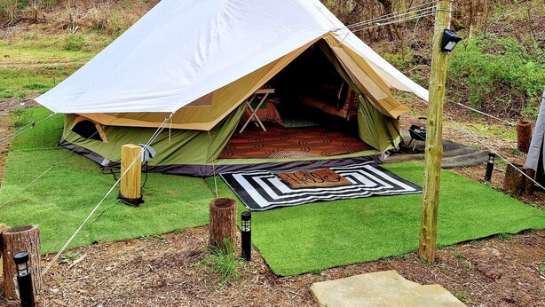 A tent with a large entrance, surrounded by green artificial grass, a patterned mat, and a wooden post.