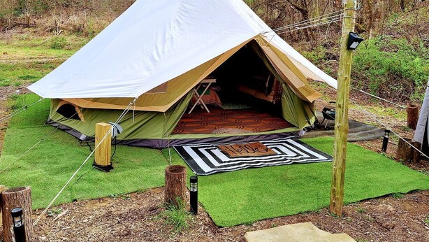A tent with a large entrance, surrounded by green artificial grass, a patterned mat, and a wooden post. - Creekside Glamping Heaven with Comfy Interior in Cosby, Tennessee (Cosby)