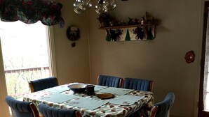 Comedor con una mesa dispuesta para cuatro personas, sillas tapizadas en azul, mantel con estampado floral y un estante decorativo en la pared.