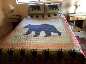 A bed with a quilt featuring a bear design, a bedside table with a lamp, and a small drawer unit.