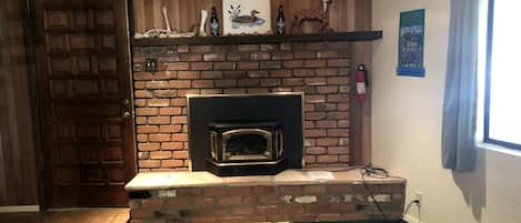 Una habitación con chimenea de ladrillo, puerta de madera, alfombra verde y ventana con cortinas.