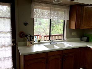 A kitchen with a double sink, wooden cabinets, a lace window valance, and a view of a deck.