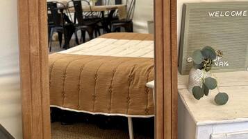 Ein Spiegel mit Holzrahmen, ein Bett mit Bettdecke, ein 'WELCOME'-Schild und ein gewebter Wandbehang.