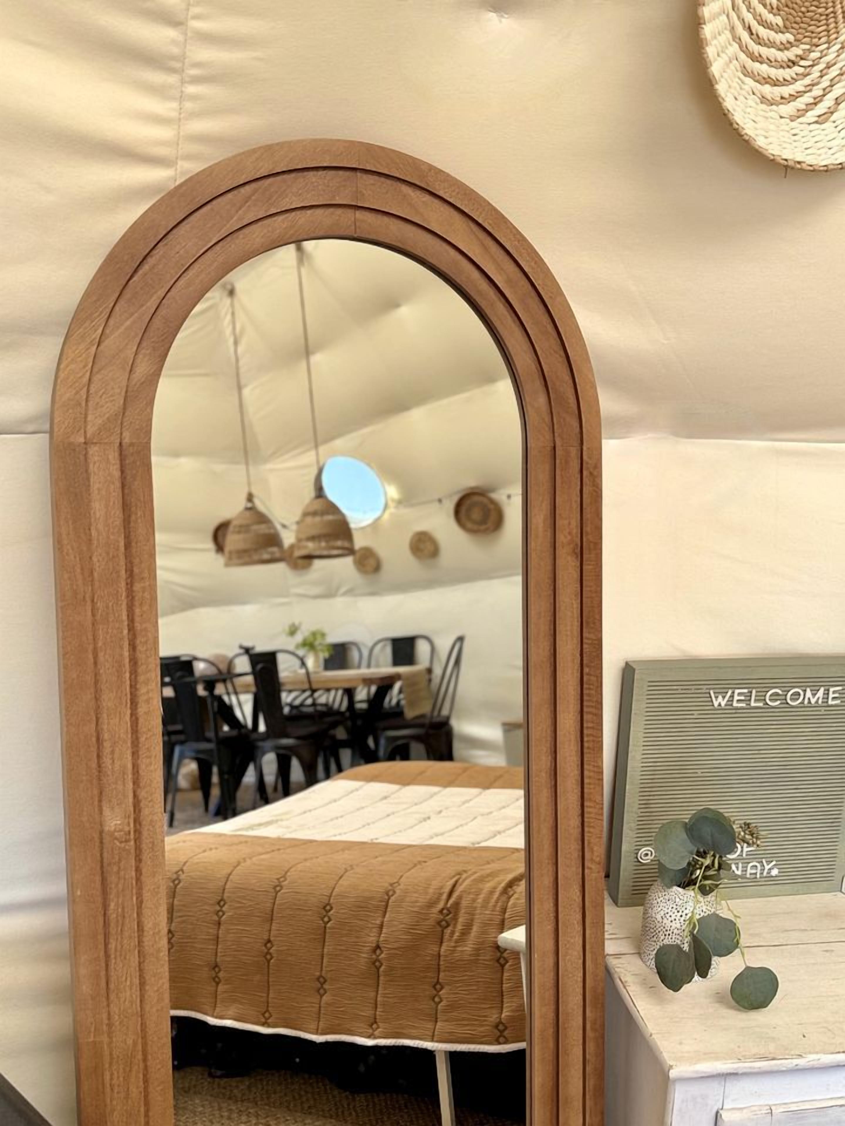 A mirror with a wooden frame, a bed with a quilt, a 'WELCOME' sign, and a woven wall hanging.